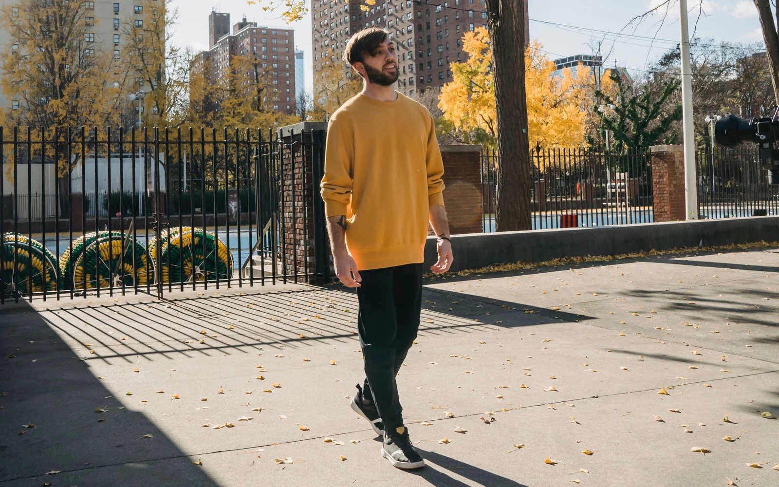 A man in a yellow sweater and full-length black trousers walks through a sunlit urban park.