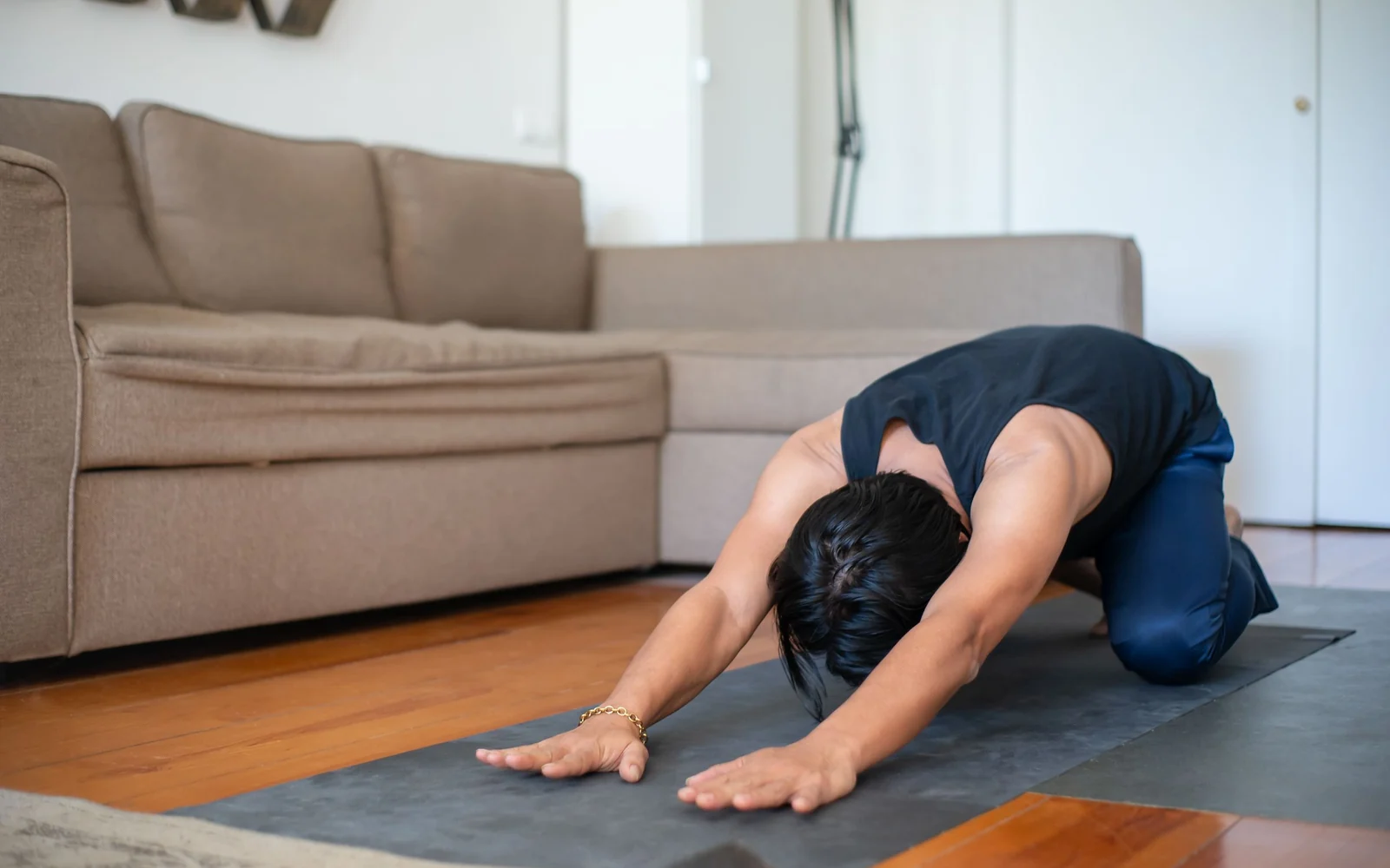 A man in full-length navy trousers reaches forward on a mat beside a sofa.