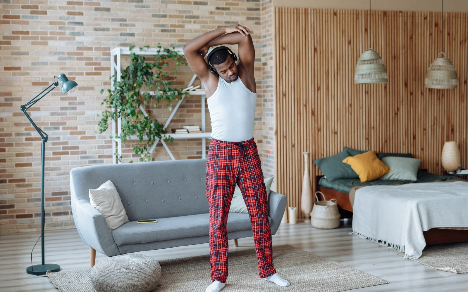 A man in full-length red checked trousers and headphones stretches his arms beside a sofa at home.