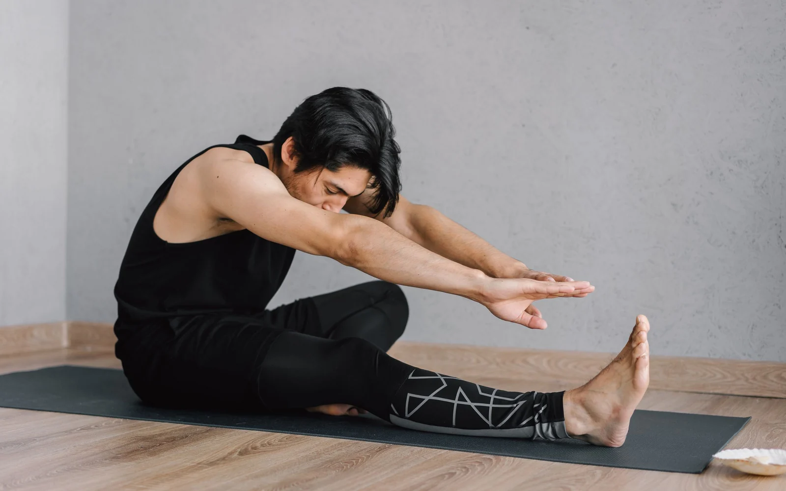 A man in full-length black trousers reaches forward over one leg on a mat.